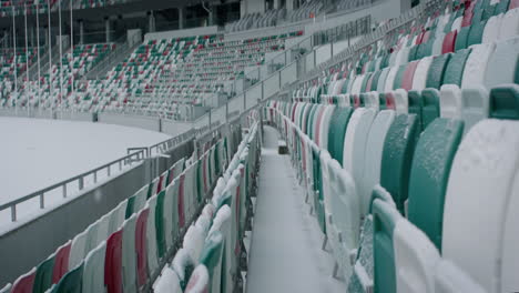 Empty-outdoor-football-soccer-stadium-seats-covered-with-snow-in-winter,-light-snowfall.-Shot-with-2x-anamorphic-lens